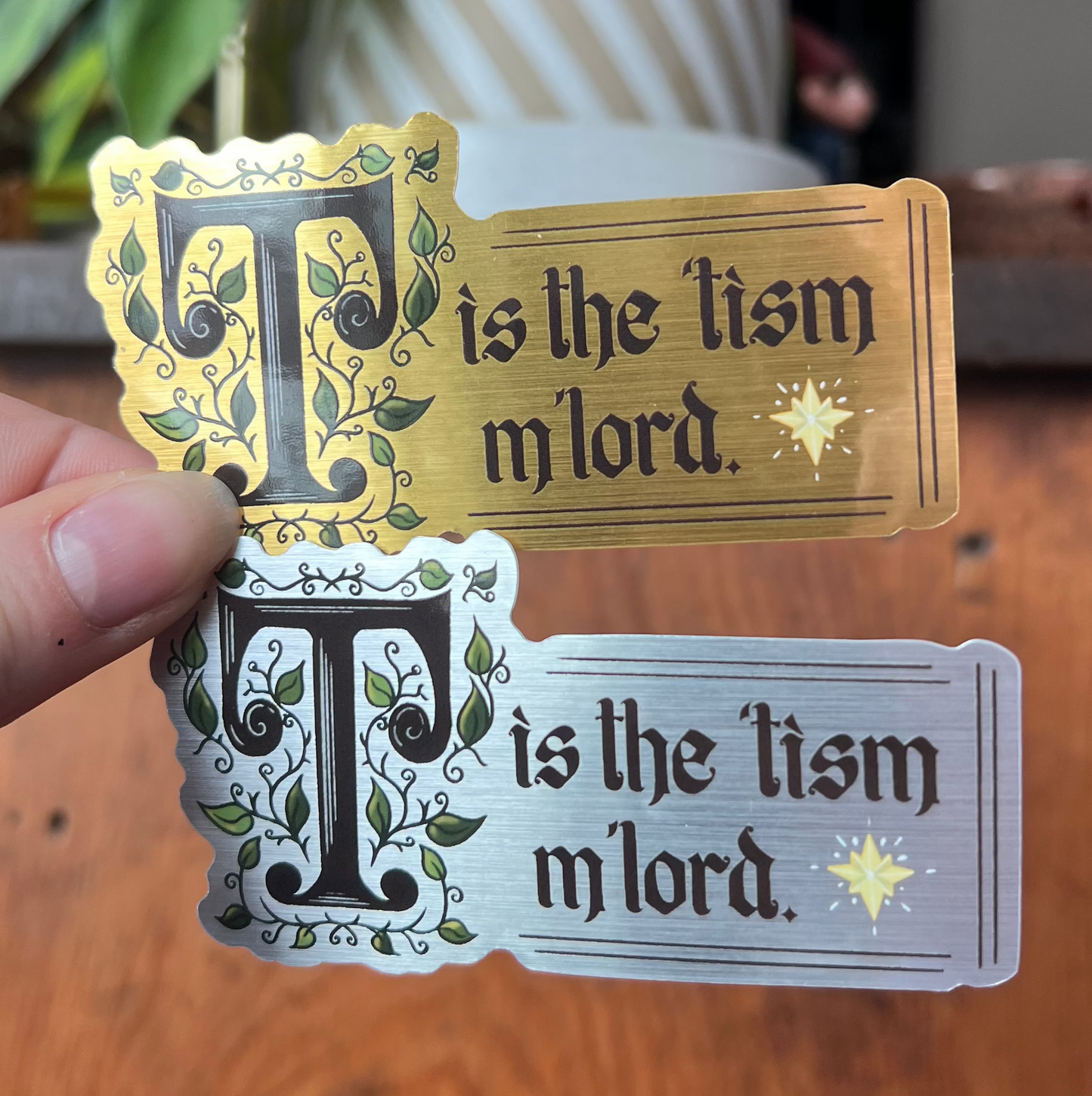Two decorative signs with 'T is the tism m'lord' text held by a hand on a wooden surface.