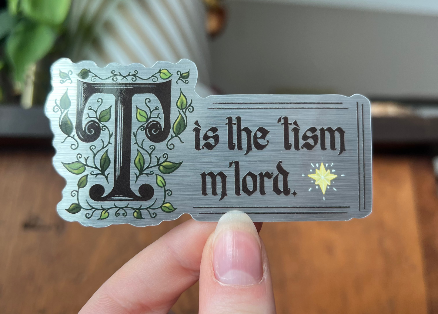 a silver decorative sticker with 'T is the tism m'lord' text held by a hand on a wooden surface.