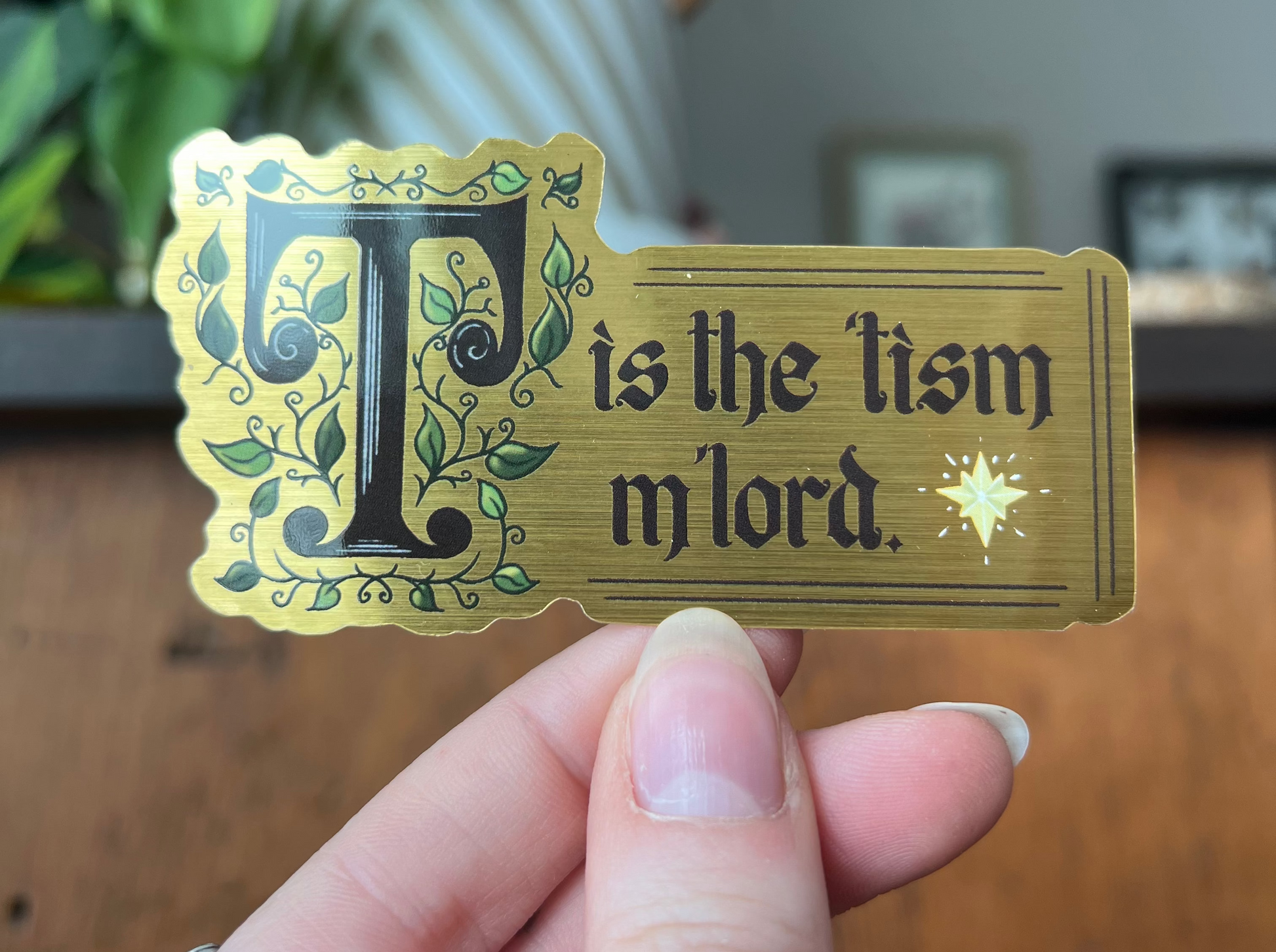 a gold decorative sticker with 'T is the tism m'lord' text held by a hand on a wooden surface.